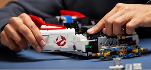 Close-up of hands assembling the LEGO Ghostbusters Ecto-1 (10274), focusing on the side panel with the iconic Ghostbusters logo and the exposed gear mechanisms during the early stages of the build.