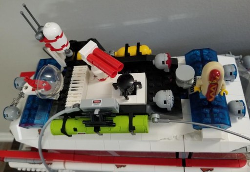 Top view of the LEGO Ghostbusters Ecto-1 roof, showcasing various intricate gadgets and equipment, including a green tank, red and white radar, and blue translucent lights. A LEGO minifigure in a hotdog costume is also visible on the roof, adding a playful touch to the detailed build.