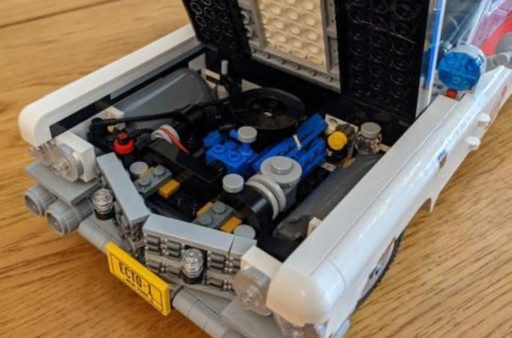 A close-up image of the LEGO Ghostbusters Ecto-1 (10274) model’s engine compartment. The hood is open, revealing a detailed engine build with various small LEGO elements representing engine components. The yellow New York license plate with “ECTO-1” is visible on the front bumper. The car’s front grille, made from intricate LEGO pieces, is also partially visible, adding to the model’s authenticity.