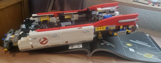 In-progress build of the LEGO Ghostbusters Ecto-1 (10274) showing the partially assembled body with visible red fins and the iconic ‘No Ghosts’ logo on the side doors. The model rests on the open instruction manual, indicating the stage of the assembly process.