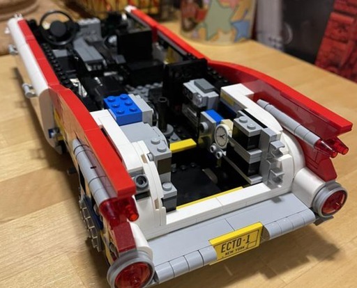 A close-up view of the rear section of the LEGO Ghostbusters Ecto-1 (10274) in progress, showing the exposed interior framework, including the rear axle and the engine compartment. The partially built model is detailed with iconic Ghostbusters symbols on the doors and a yellow “ECTO-1” New York license plate on the back bumper. The rear fins and tail lights of the car are also visible in the image.