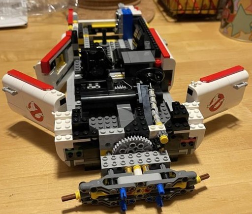 A partially constructed LEGO Ghostbusters Ecto-1 (10274) model. The image shows the Technic frame of the vehicle in progress, with the car doors open, revealing the interior steering mechanism and early stages of the gear assembly. This frame is crucial for providing structural support as the build progresses. The Ghostbusters logo is visible on the car doors, and various gears and Technic components are in place, which will later enable the interactive features of the model. The scene is set on a wooden table, highlighting the build in a relaxed, home environment.