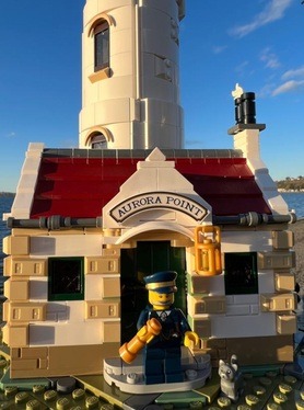 Close up image of the Lego motorized Lighthouse outdoors, of the entrance to the lighthouse with the lighthouse keeper in front 